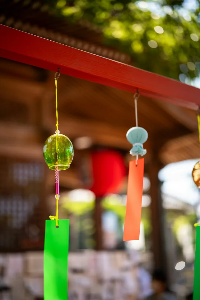 Rows of Japanese glass fūrin wind chimes with paper strips hang beneath a red structure against summer greenery
