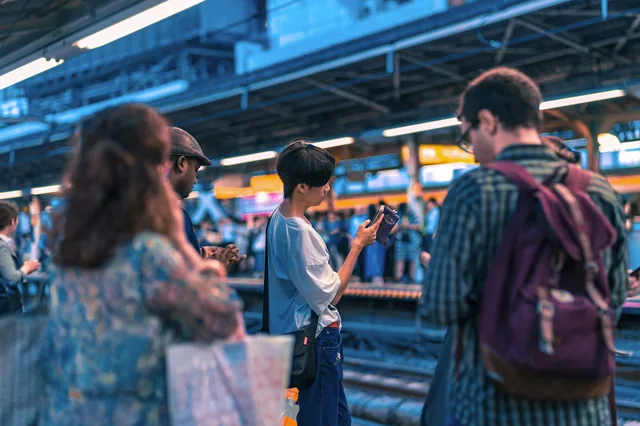 Travellers wait for a train at Harajuku Station in Tokyo while one checks his phone near the yellow safety line
